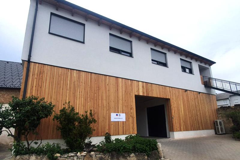 Modernes zweistöckiges Gebäude mit Holzverkleidung, drei Fenstern und einer offenen Garageneinfahrt im Erdgeschoss.