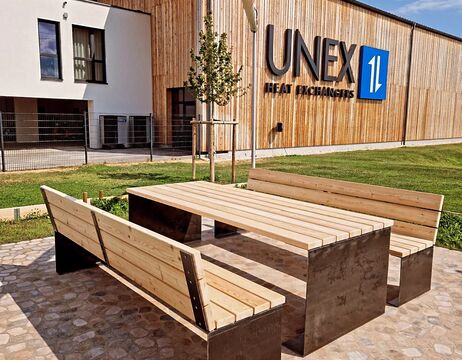 Holzbänke und Tisch im Freien auf einer Steinterrasse vor einem Gebäude mit der Aufschrift UNEX Heat Exchangers.