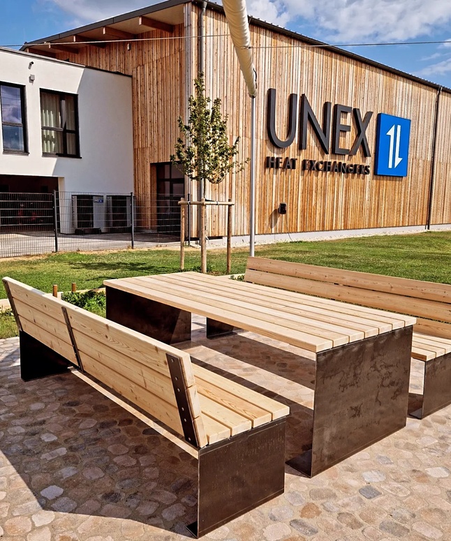 Holzbänke und Tisch im Freien auf einer Steinterrasse vor einem Gebäude mit der Aufschrift UNEX Heat Exchangers.