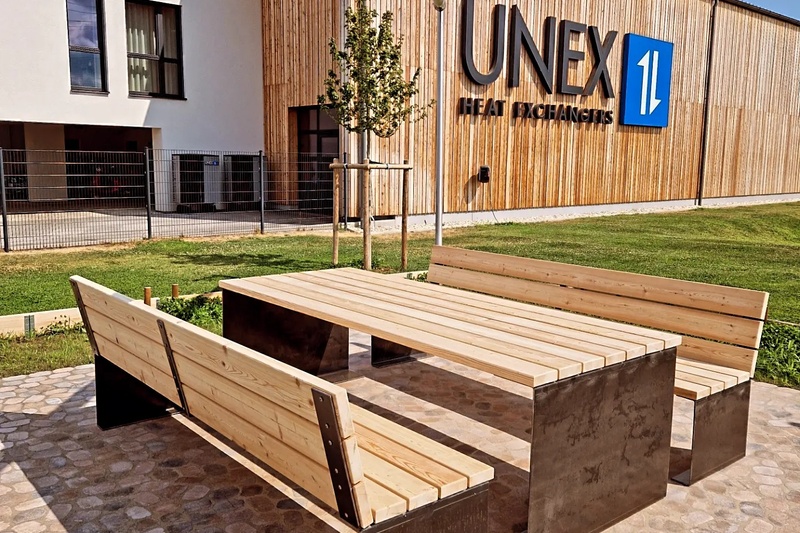 Holzbänke und Tisch im Freien auf einer Steinterrasse vor einem Gebäude mit der Aufschrift UNEX Heat Exchangers.