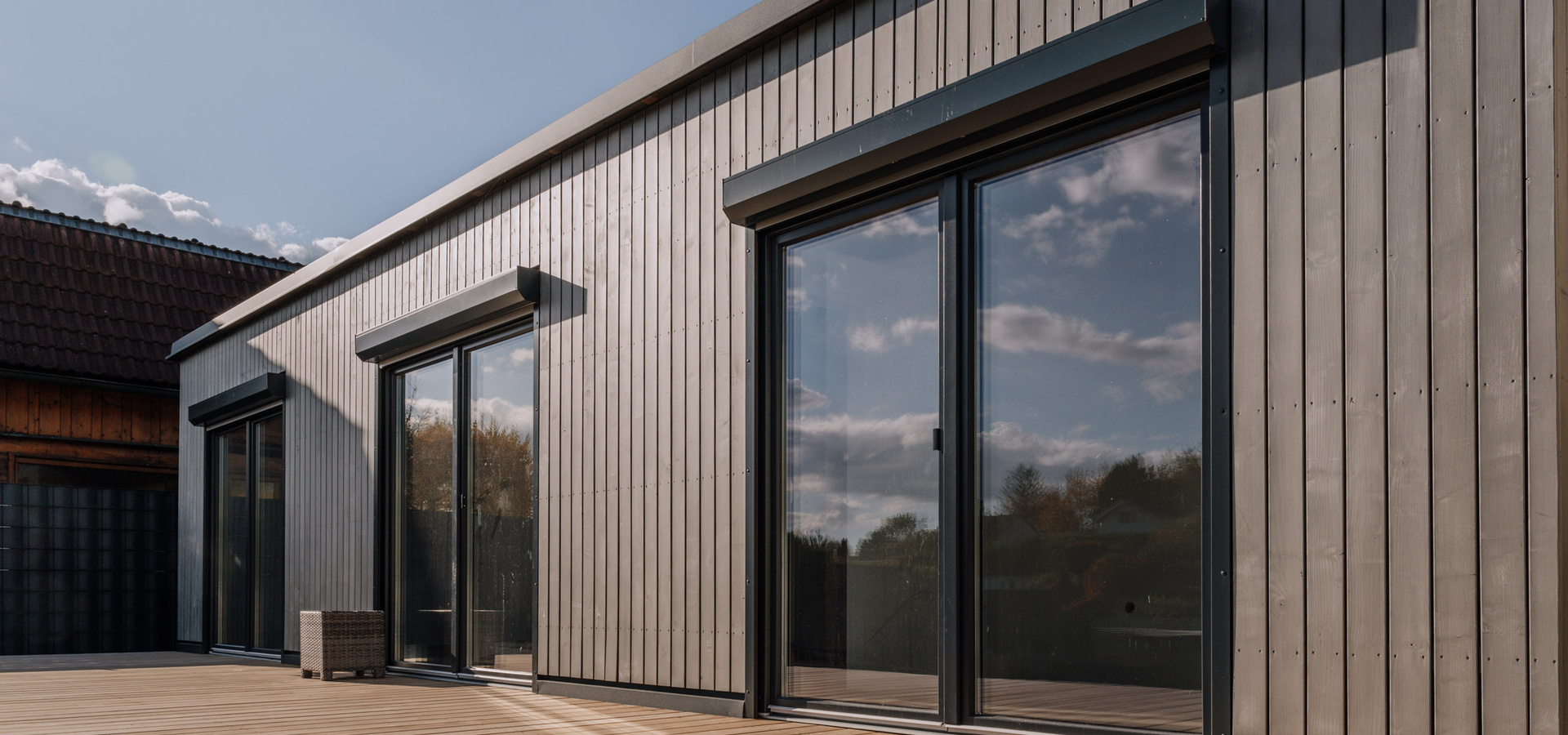 Modernes Holzhaus mit großen Glasschiebetüren, in denen sich Himmel und Wolken spiegeln, und einem kleinen Stuhl im Freien.