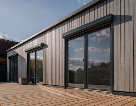 Modernes Holzhaus mit großen Glasschiebetüren, in denen sich Himmel und Wolken spiegeln, und einem kleinen Stuhl im Freien.