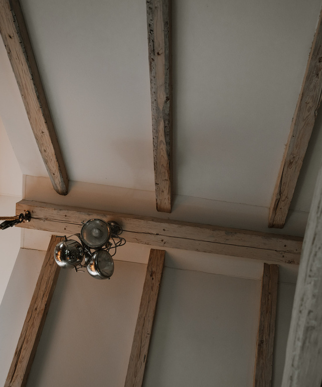 Holzbalken an der Decke und ein Metallkronleuchter, von unten betrachtet, in einem sanft beleuchteten Raum.