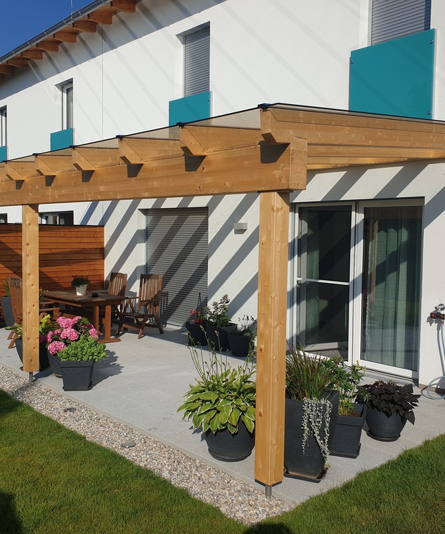 Modernes Haus mit einer hölzernen Pergola über einem Innenhof, umgeben von Topfpflanzen, Stühlen und einem Tisch.