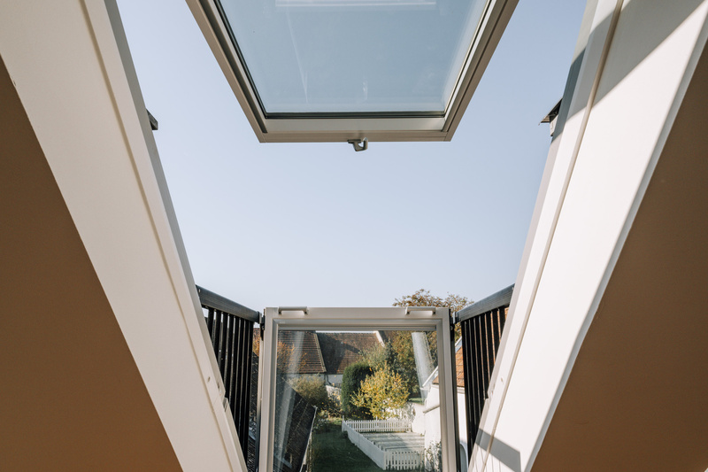 Offenes Oberlichtfenster über einer Treppe mit Blick auf die Dächer und einen Garten mit weißem Zaun.