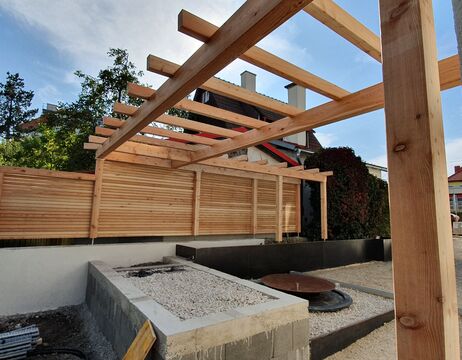 Im Bau befindliche Pergolastruktur aus Holz in einem Garten mit Kies- und Betonpflanzbeeten.
