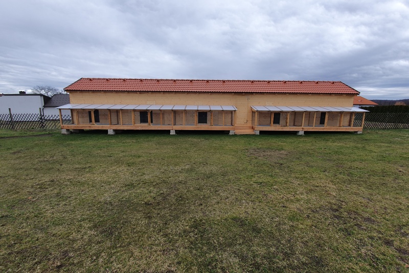 Ein langes, gelbes Gebäude mit kleinen überdachten Fenstern steht auf einem grasbewachsenen Rasen unter einem bewölkten Himmel.