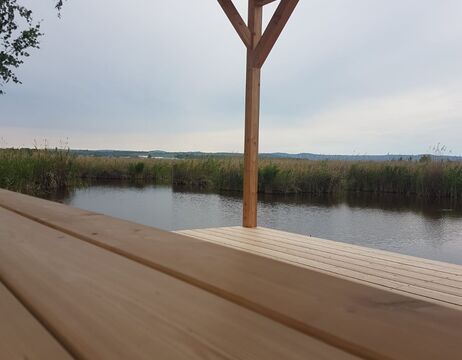 Holzsteg mit Vordach und Blick auf einen ruhigen Fluss und einen grasbewachsenen Sumpf bei bewölktem Himmel.