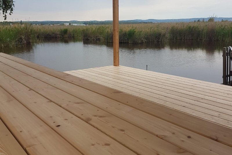 Ein Holzdeck überragt ein ruhiges Gewässer, umgeben von Schilf und fernen Hügeln unter einem bewölkten Himmel.