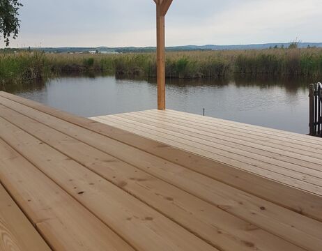 Ein Holzdeck überragt ein ruhiges Gewässer, umgeben von Schilf und fernen Hügeln unter einem bewölkten Himmel.