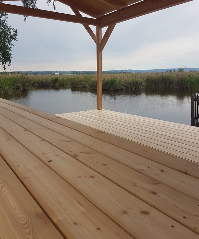 Ein Holzdeck überragt ein ruhiges Gewässer, umgeben von Schilf und fernen Hügeln unter einem bewölkten Himmel.