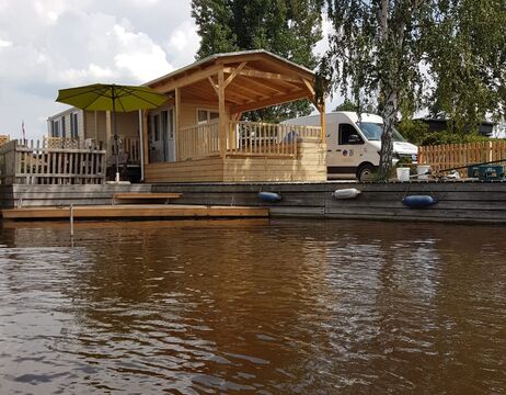 Holzhütte mit Veranda und grünem Sonnenschirm an einem Fluss, in dessen Nähe ein weißer Lieferwagen parkt.