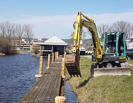 Ein kleiner Bagger repariert an einem klaren, sonnigen Tag einen hölzernen Steg an einem Fluss.