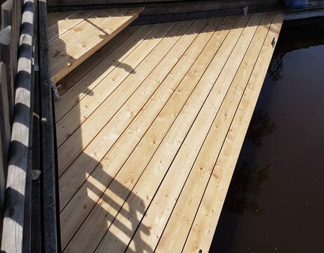 Hölzerner Steg mit Sonnenlicht, das den Schatten eines Stuhls am dunklen Wasser wirft.