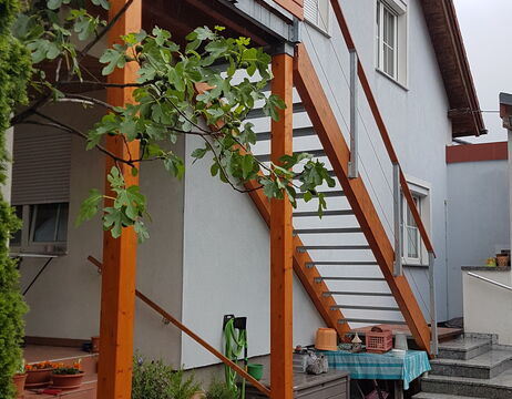 Außentreppe aus Holz mit Metallgeländer an einem weißen Haus, neben einem grünen, belaubten Baum.