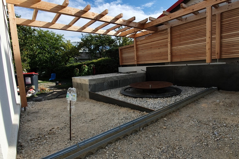 Außenterrasse unter hölzerner Pergola, mit Kiesboden und teilweise aufgebautem Sitzbereich.