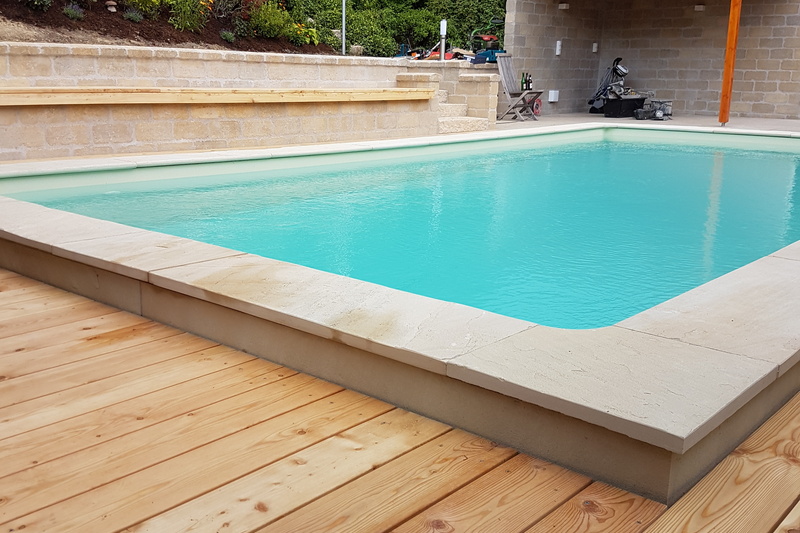 Rechteckiger Swimmingpool mit klarem Wasser, Steinrand und Holzdeck neben einer Backsteinmauer in einem Hinterhof.