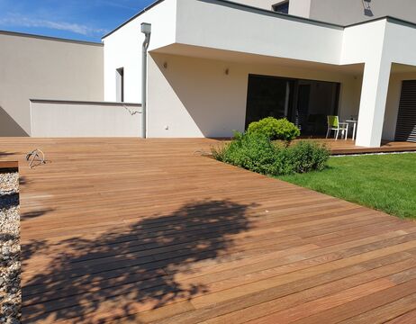 Moderner Innenhof mit Holzterrasse, grünen Büschen und einer kleinen Rasenfläche unter sonnigem Himmel.