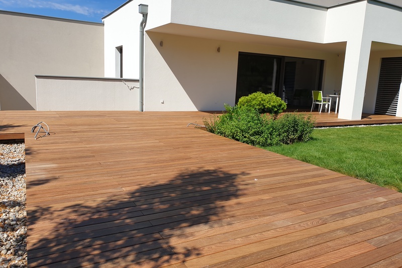 Moderner Innenhof mit Holzterrasse, grünen Büschen und einer kleinen Rasenfläche unter sonnigem Himmel.