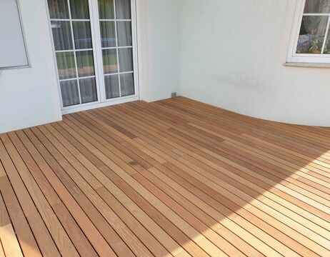 Ein Holzdeck mit hellen Dielen neben einer weißen Wand und Terrassentüren aus Glas.