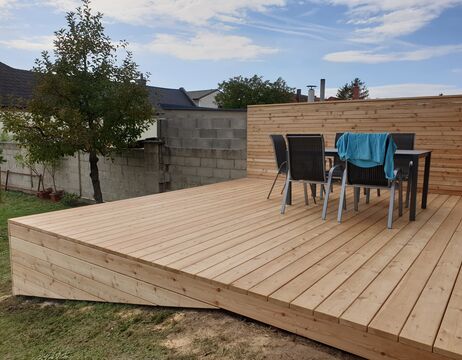Moderne Holzterrasse mit einem Tisch, vier Stühlen und einem blauen Tuch, das über einen Stuhl drapiert ist, in einem Hinterhof.