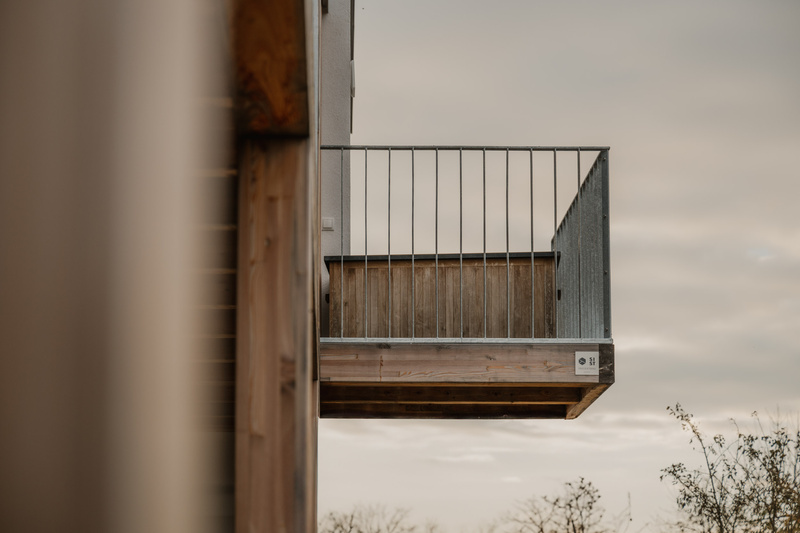 Ein moderner, an einem Gebäude angebrachter Balkon aus Holz und Metall, von der Seite gegen einen bewölkten Himmel fotografiert.
