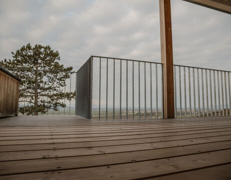 Eine Holzterrasse mit Metallgeländer bietet einen Blick auf eine malerische, wolkenverhangene Landschaft und ferne Bäume.
