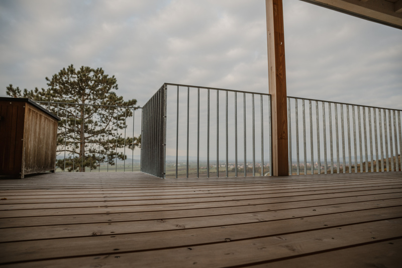 Eine Holzterrasse mit Metallgeländer bietet einen Blick auf eine malerische, wolkenverhangene Landschaft und ferne Bäume.