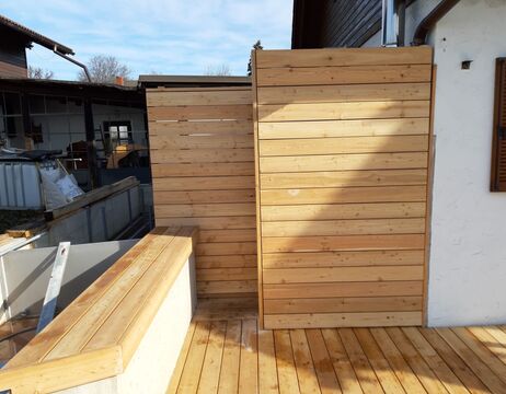 Sichtschutz aus Holz und Bank auf einer Holzterrasse neben einem Haus in einem Hinterhof.