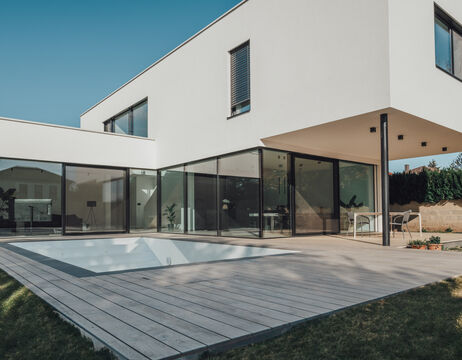 Modernes weißes Haus mit großen Glasfenstern und einem leeren Pool auf einem Holzdeck im Hinterhof.