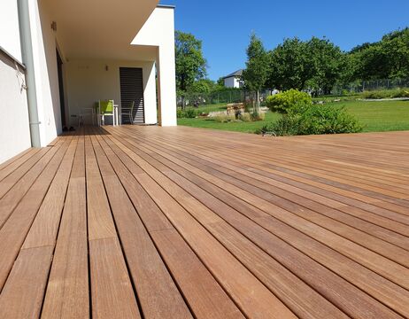 Die Holzterrasse erstreckt sich über ein modernes Haus und bietet einen Blick auf einen grünen Garten mit Bäumen unter einem blauen Himmel.