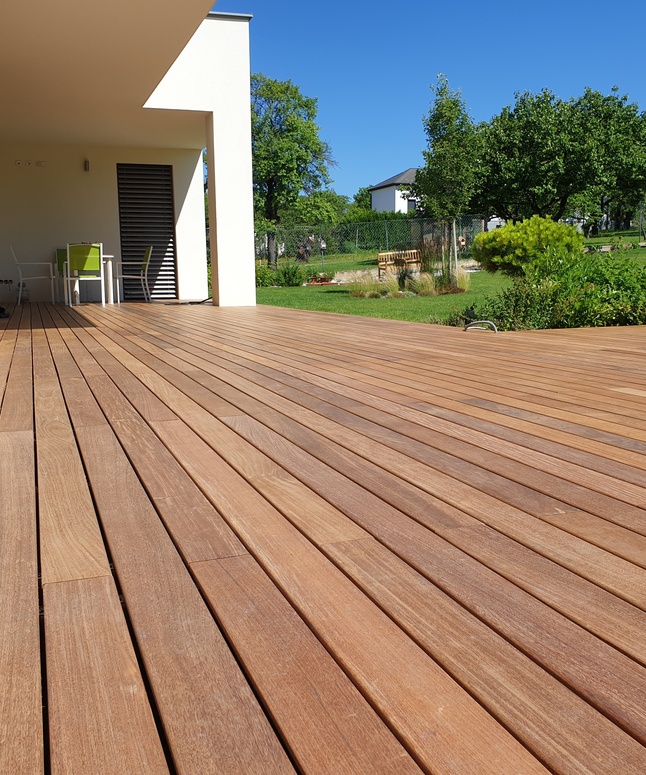 Die Holzterrasse erstreckt sich über ein modernes Haus und bietet einen Blick auf einen grünen Garten mit Bäumen unter einem blauen Himmel.