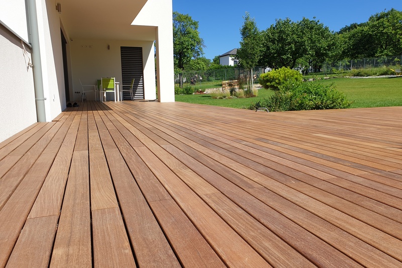 Die Holzterrasse erstreckt sich über ein modernes Haus und bietet einen Blick auf einen grünen Garten mit Bäumen unter einem blauen Himmel.