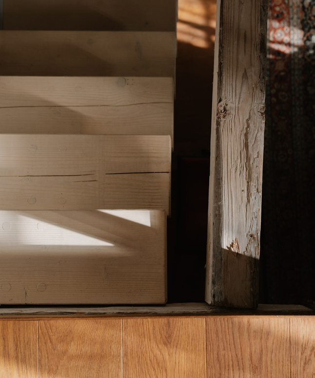 Das Sonnenlicht wirft Schatten auf eine helle Holztreppe, die von oben gesehen neben einem verwitterten Holzpfosten steht.
