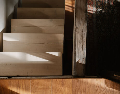 Das Sonnenlicht wirft Schatten auf eine helle Holztreppe, die von oben gesehen neben einem verwitterten Holzpfosten steht.