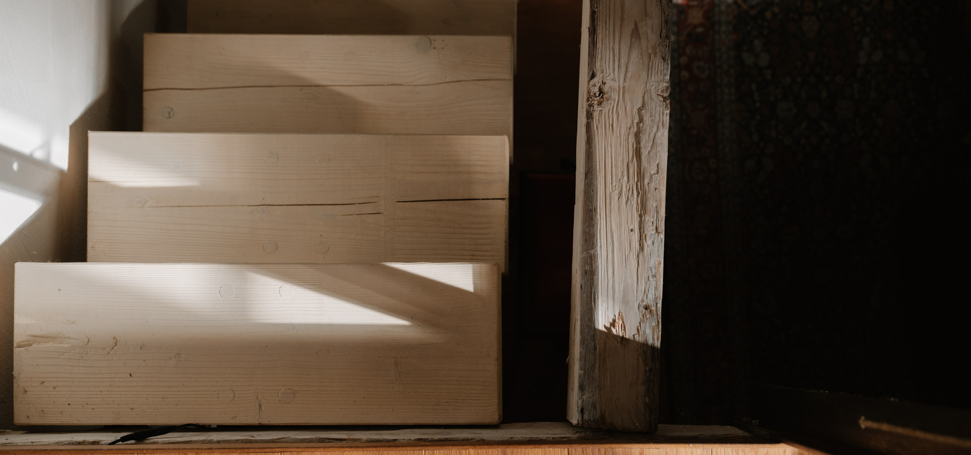 Das Sonnenlicht wirft Schatten auf eine helle Holztreppe, die von oben gesehen neben einem verwitterten Holzpfosten steht.