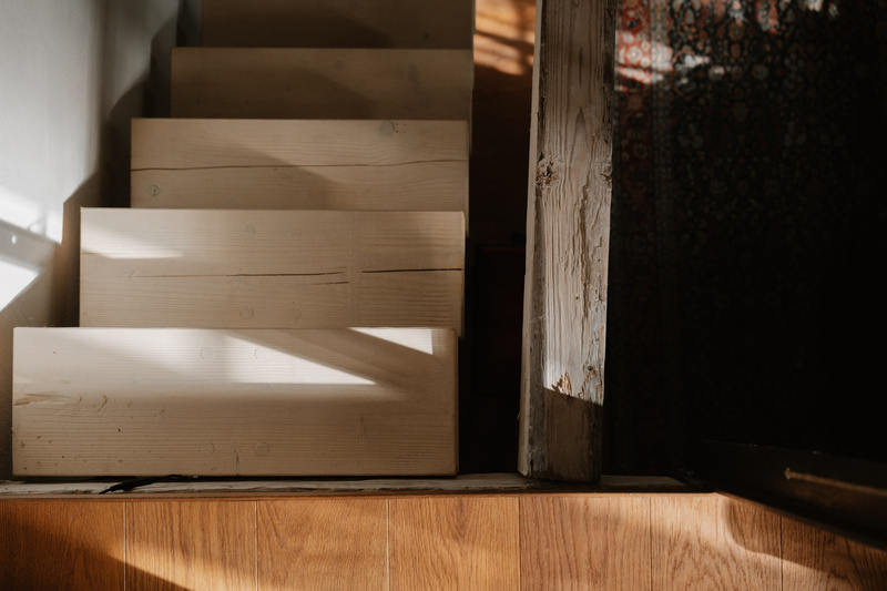 Das Sonnenlicht wirft Schatten auf eine helle Holztreppe, die von oben gesehen neben einem verwitterten Holzpfosten steht.