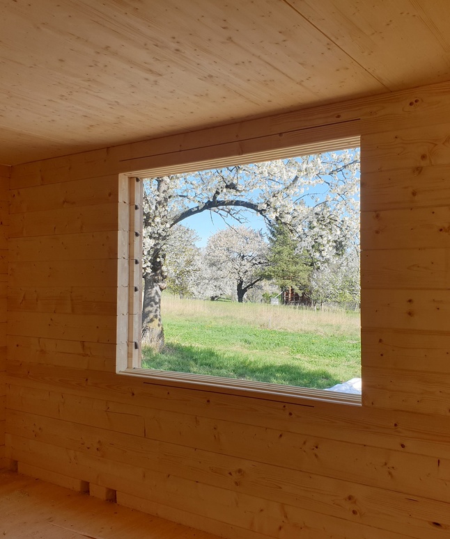 Ein hölzernes Zimmer mit einem Fenster, das blühende Bäume und grünes Gras an einem sonnigen Tag zeigt.