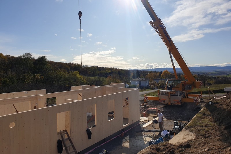 Ein Kran hebt Materialien auf einer Baustelle mit teilweise errichteten Holzwänden unter einem sonnigen Himmel.