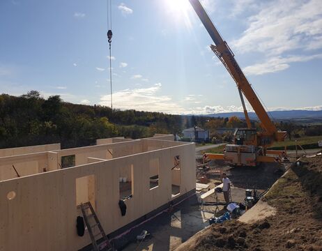 Ein Kran hebt Materialien auf einer Baustelle mit teilweise errichteten Holzwänden unter einem sonnigen Himmel.