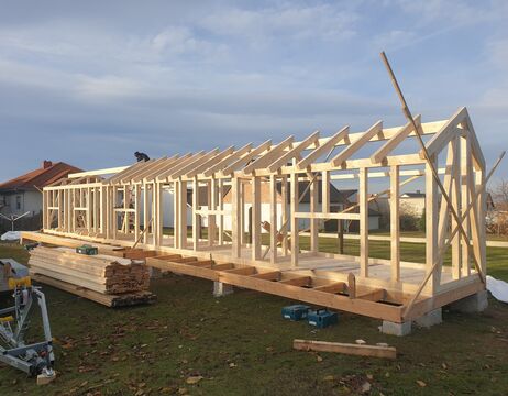 Im Bau befindliches Holzhaus auf einem grasbewachsenen Grundstück mit Bauholz und Werkzeugen in der Nähe.