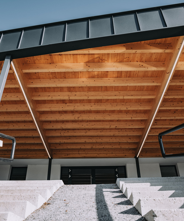 Blick von unten auf eine moderne Holzdachkonstruktion über einer Betontreppe mit schwarzem Geländer.