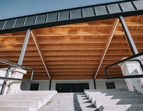 Blick von unten auf eine moderne Holzdachkonstruktion über einer Betontreppe mit schwarzem Geländer.
