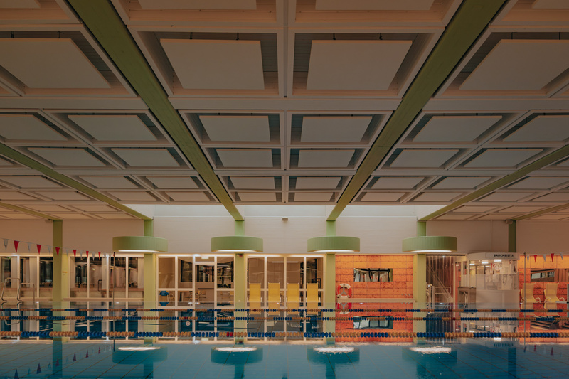 Hallenbad mit Bahnen, bunten Fahnen und Sitzgelegenheiten unter einer gitterartig gestalteten Decke.