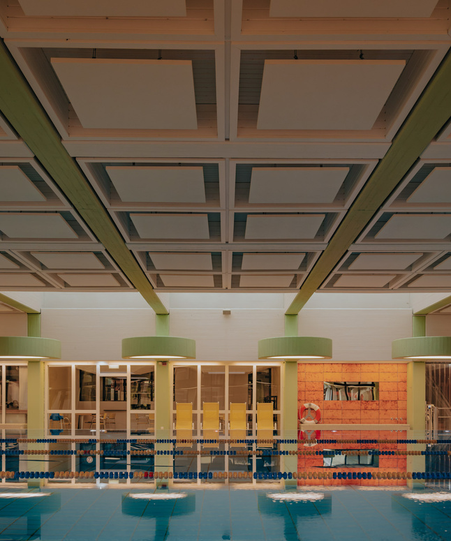 Hallenbad mit Bahnen, bunten Fahnen und Sitzgelegenheiten unter einer gitterartig gestalteten Decke.