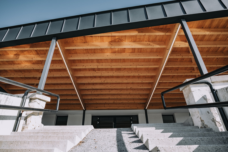 Eine breite Treppe führt zu einem modernen Gebäude mit freiliegenden Holzbalken und einer schwarzen Metallbedachung.