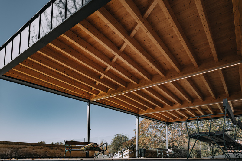 Terrassenüberdachung aus Holz mit freiliegenden Balken, Metallstützen und Gartenmöbeln darunter.