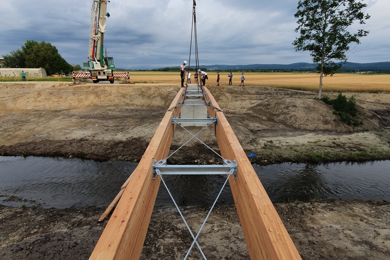 Auf der Baustelle wird eine hölzerne Fußgängerbrücke über einen Bach installiert, wobei Menschen und ein Kran im Einsatz sind.