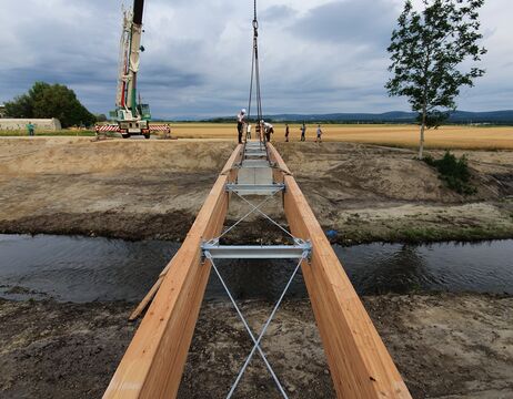 Auf der Baustelle wird eine hölzerne Fußgängerbrücke über einen Bach installiert, wobei Menschen und ein Kran im Einsatz sind.