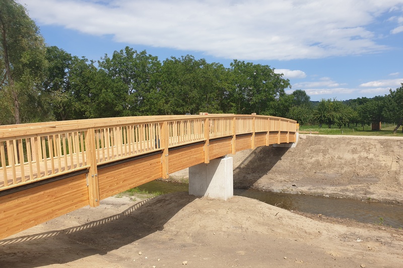 Eine hölzerne Fußgängerbrücke überspannt einen kleinen Bach, umgeben von Bäumen und einem klaren Himmel.